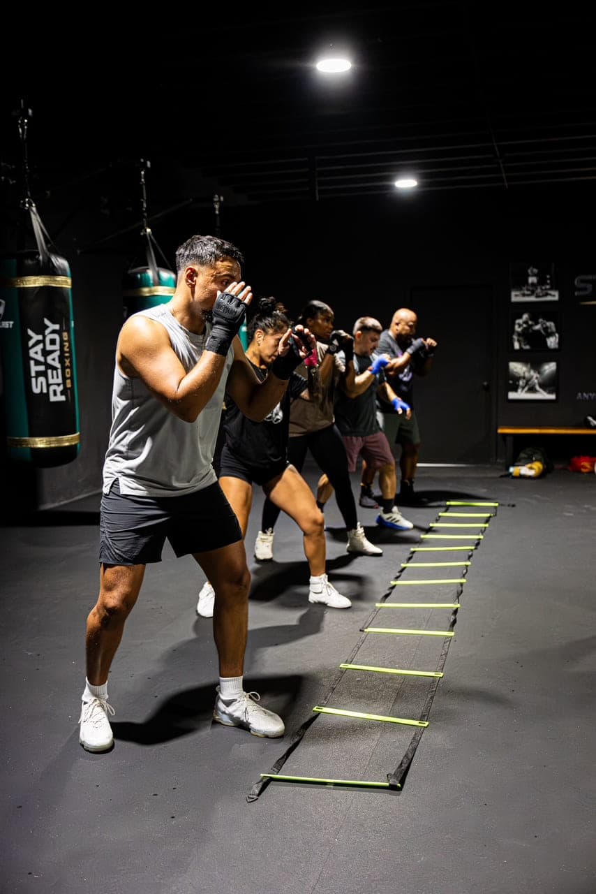 A line of members shadowboxing in guard beside an agility ladder, Stay Ready Boxing Gym heavy bags hanging to the left.