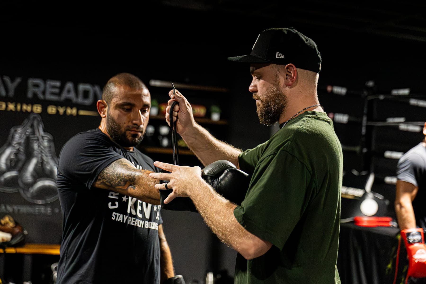 Coach Kevin Gleason fastening a boxing glove onto a member’s hand on the training floor, the Stay Ready Boxing Gym mural on the wall behind them.