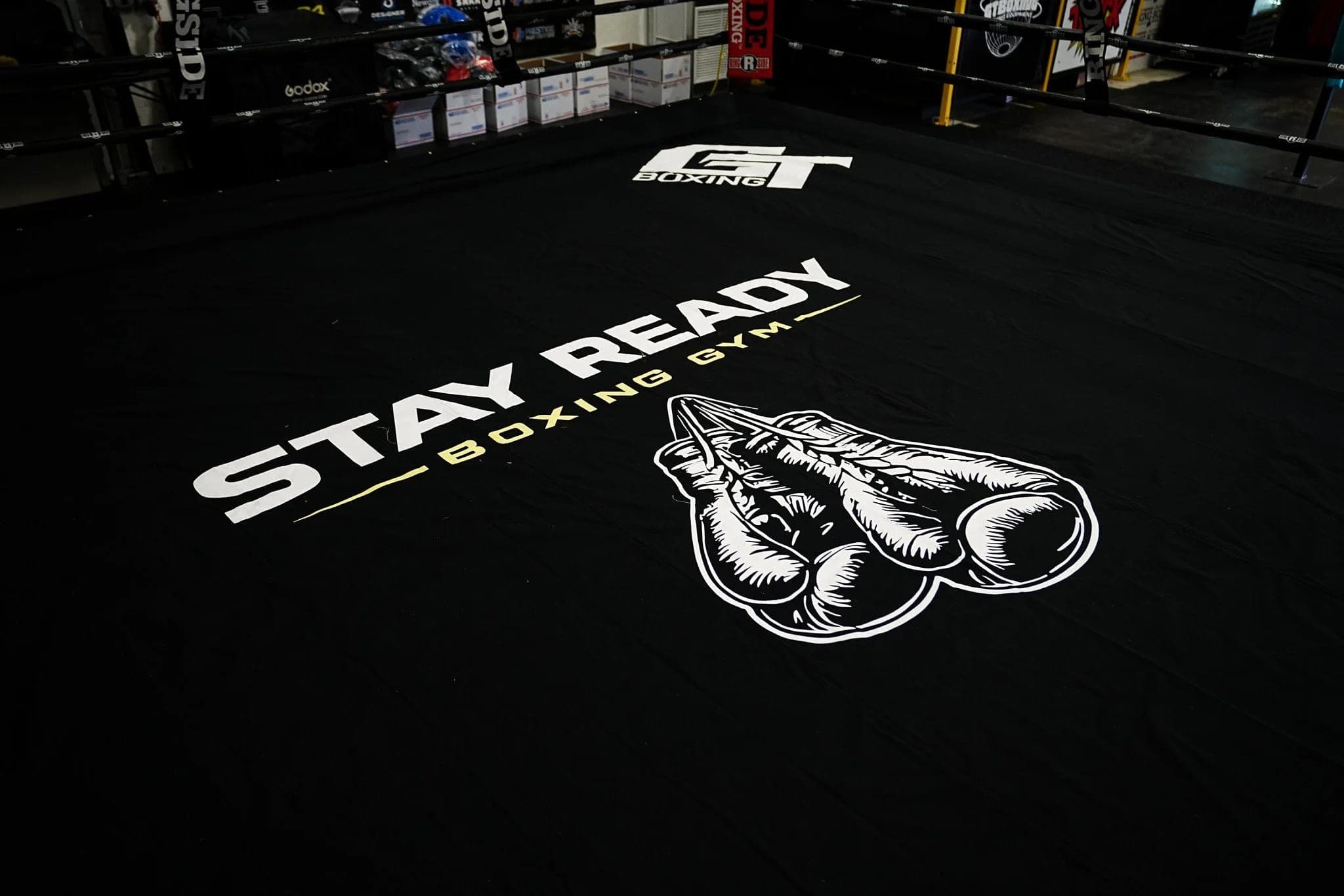 The Stay Ready Boxing Gym ring seen from a corner, the gym logo printed across the black canvas, ropes running across the frame.
