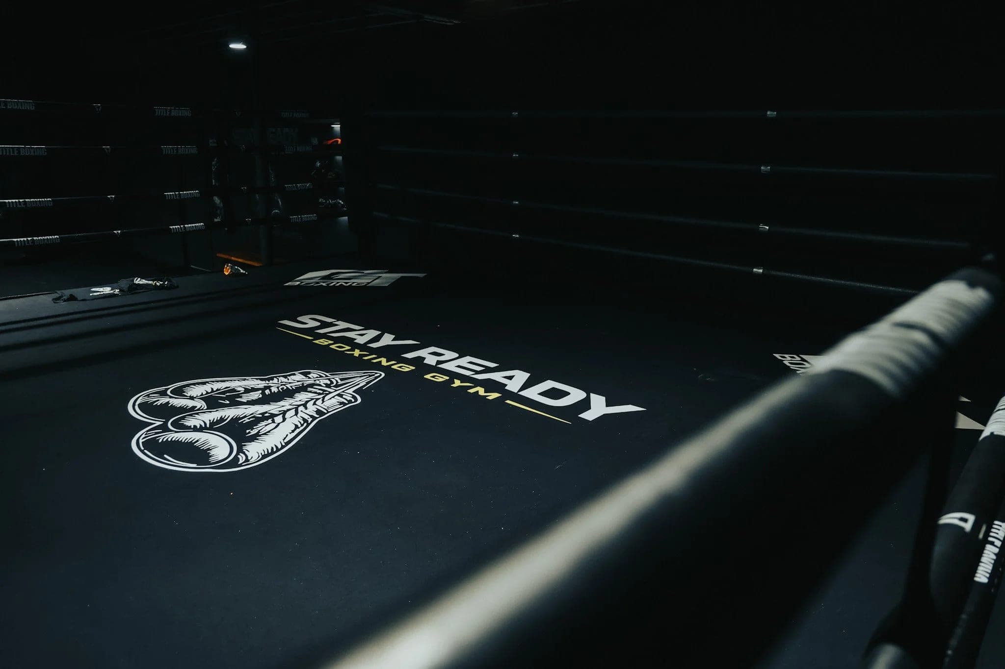 The Stay Ready Boxing Gym ring under low light, a taped rope running diagonally across the foreground and the gym logo on the canvas beyond it.