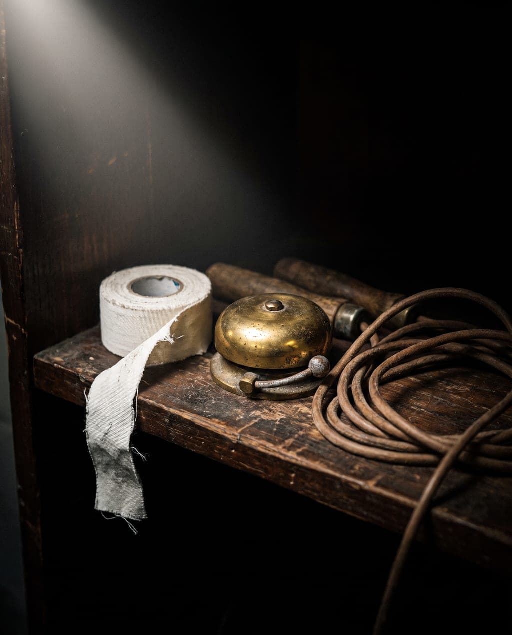 A roll of white athletic tape, a brass round-timer bell and a coiled jump rope on a dark wooden shelf.